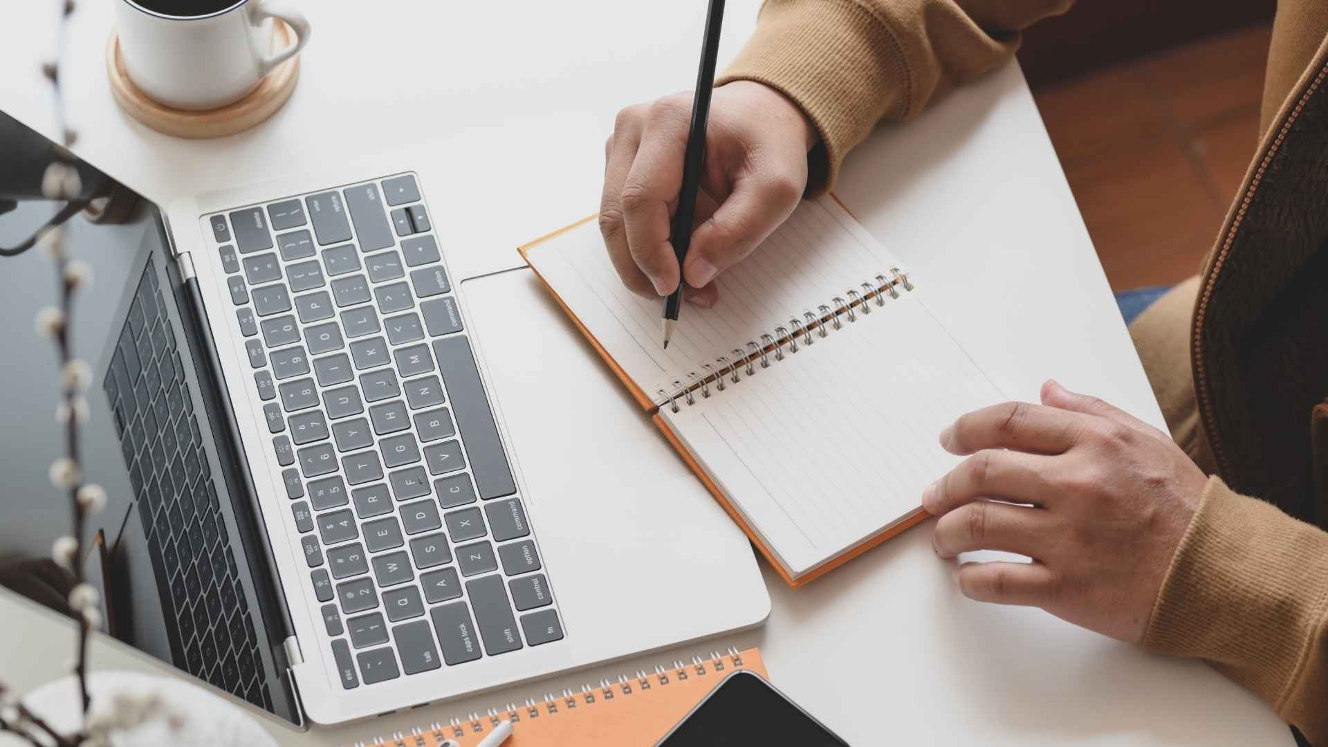 person writing on notebook next to laptop