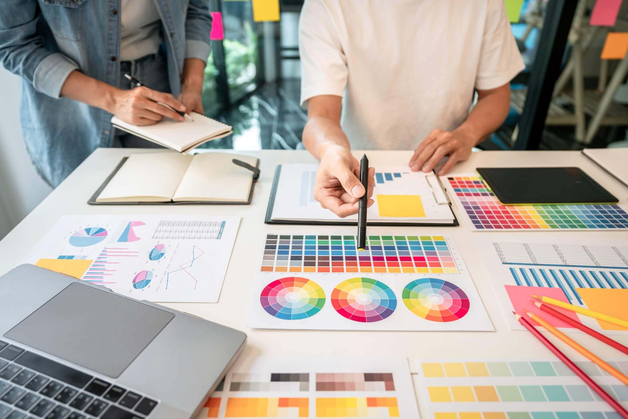 Creative professionals discussing over a table scattered with color swatches, graphs, and design mockups, indicating a collaborative graphic design project.