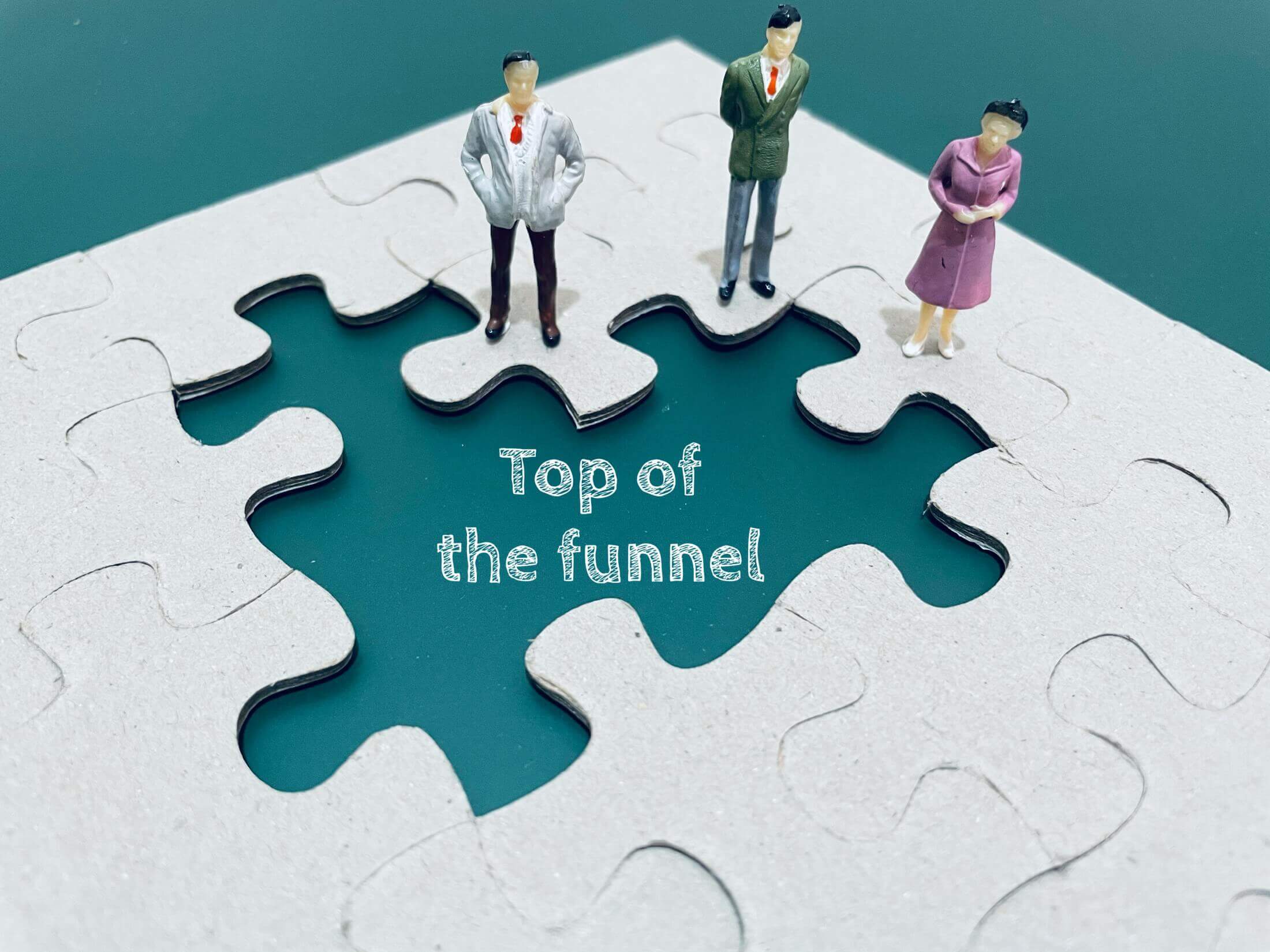 An image depicting a puzzle with missing pieces, where the visible pieces are fitted with figurines standing on them, and the phrase "Top of the funnel" is displayed, symbolizing a marketing concept.
